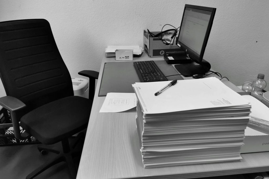 Black and white image of a cluttered office desk with a computer, chair, and paperwork.