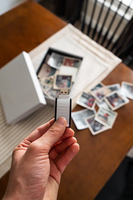 Hand holding a USB drive above a table with scattered vintage photos and an open photo box, symbolizing digital archiving.